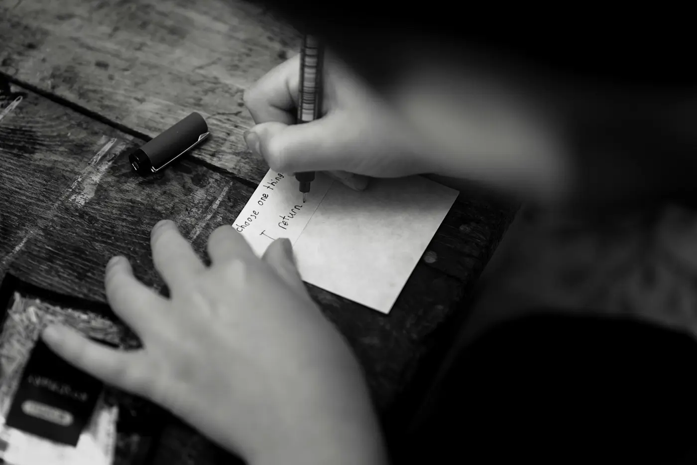 A hand writing a short note on a small card at a weathered wooden desk.