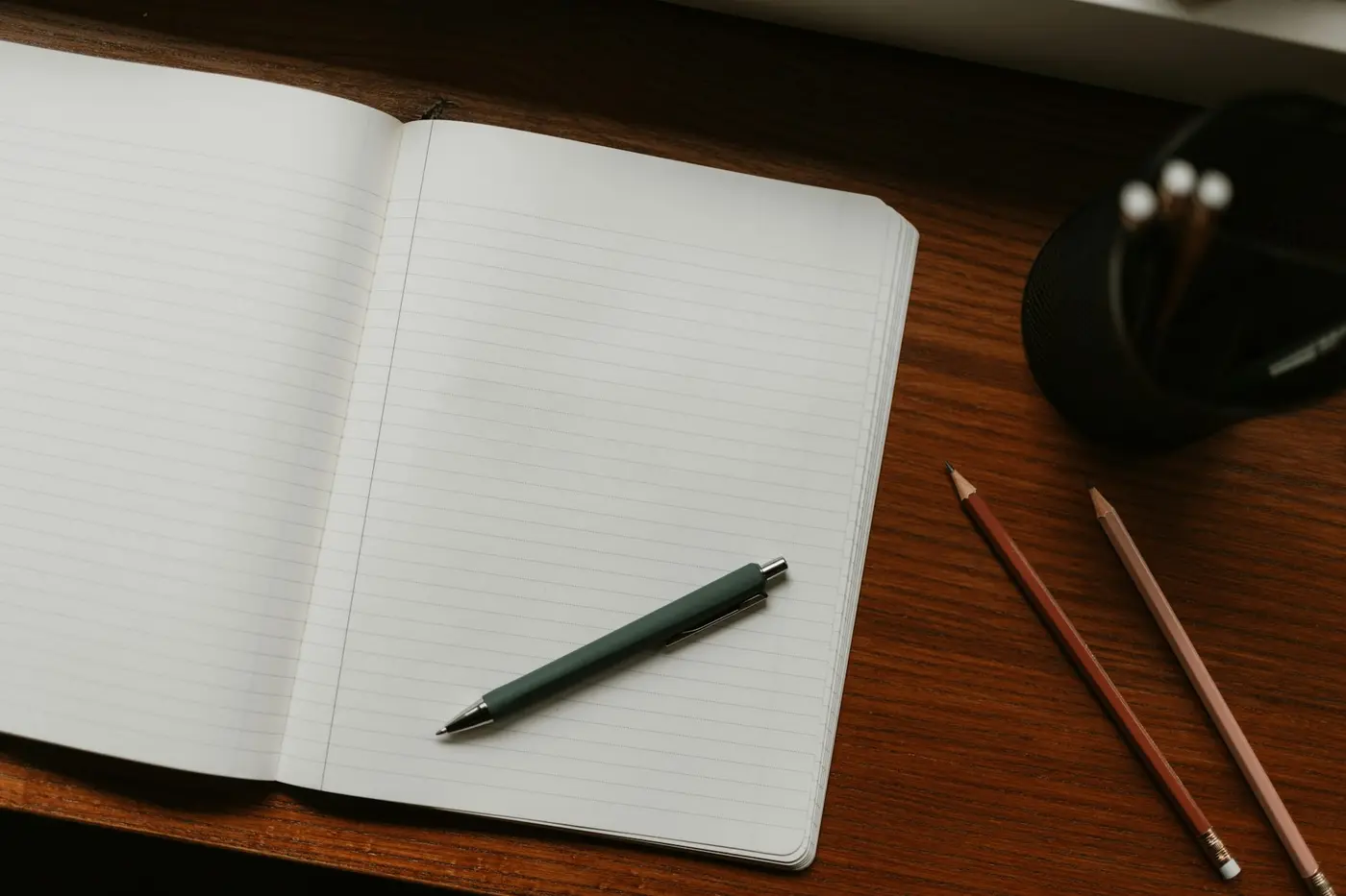 An open ruled notebook and pen on a dark wooden desk, with two pencils and a pen pot beside it.