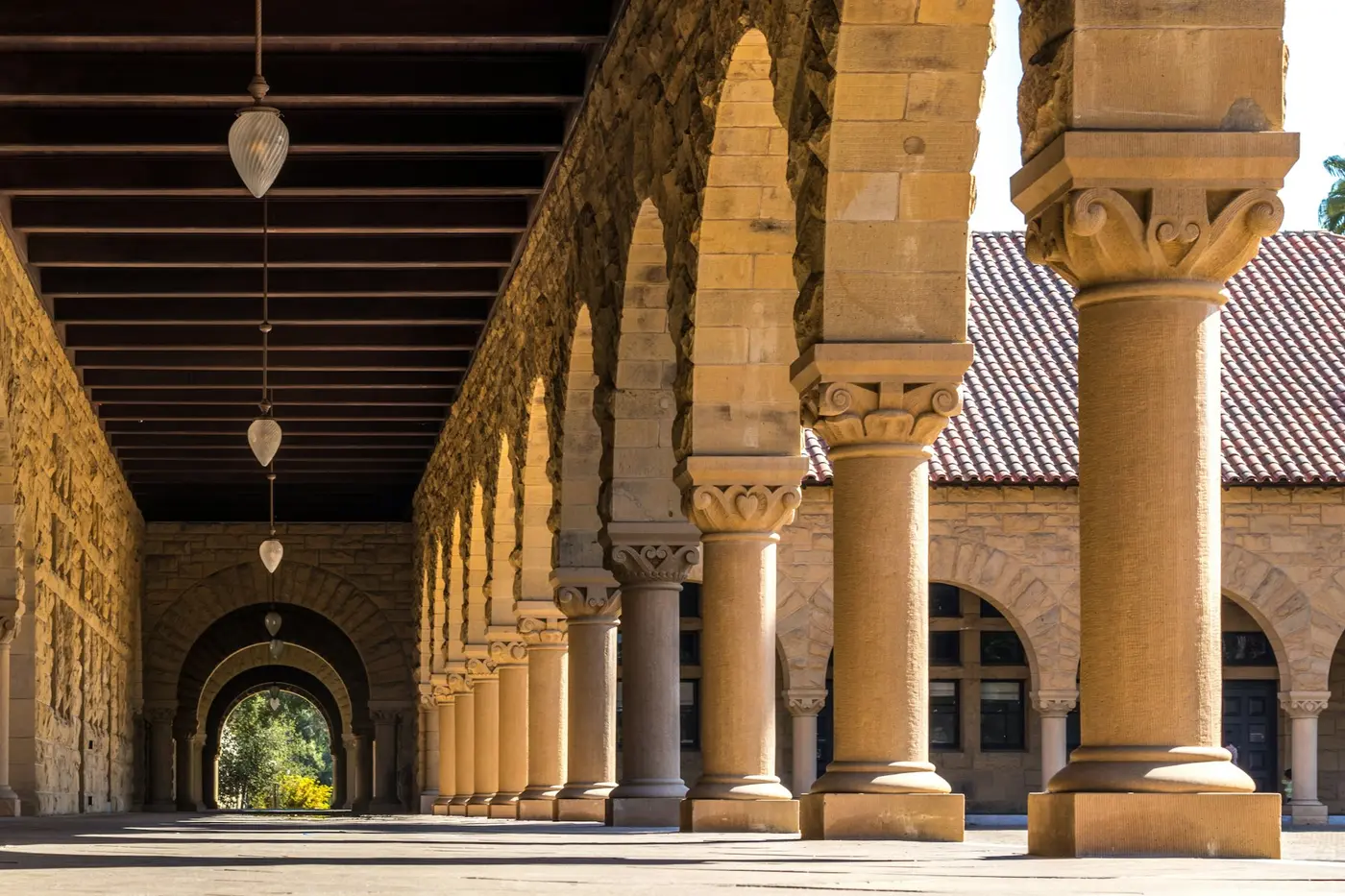 A sandstone arcade of carved columns and round arches, with lamps hanging from a timber ceiling.
